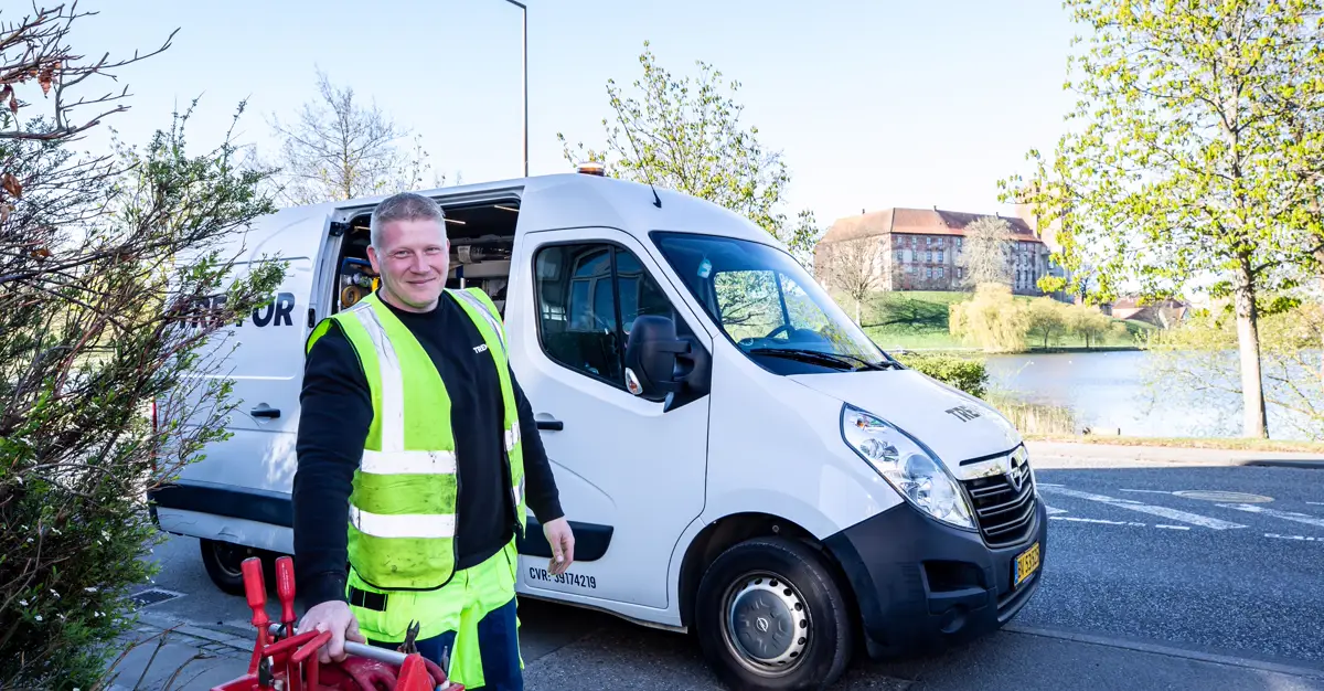A tradesperson in a reflective vest standing beside their work van holding tools.