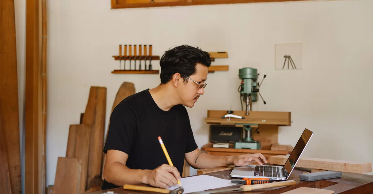A carpenter planning a woodworking project on a laptop in their workshop.