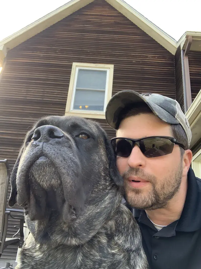 Man in camo cap and sunglasses posing close to a large gray mastiff outside a brown house