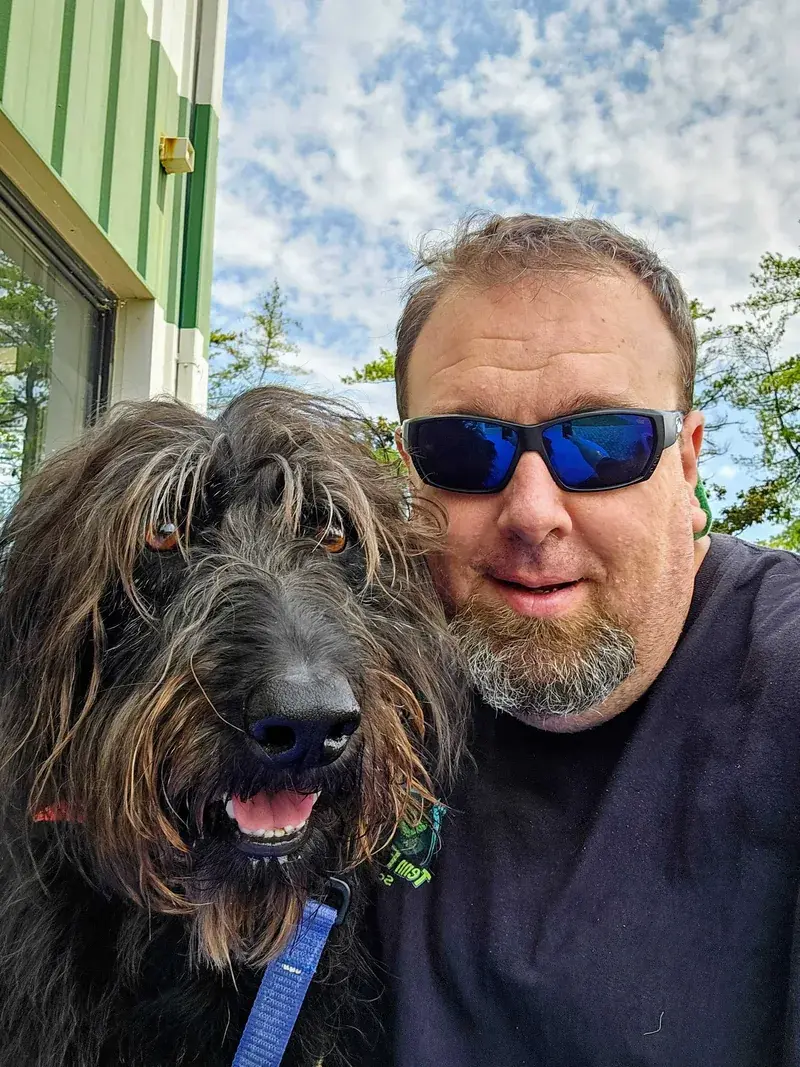 Smiling bearded man in blue sunglasses beside a shaggy dark dog under a cloudy blue sky